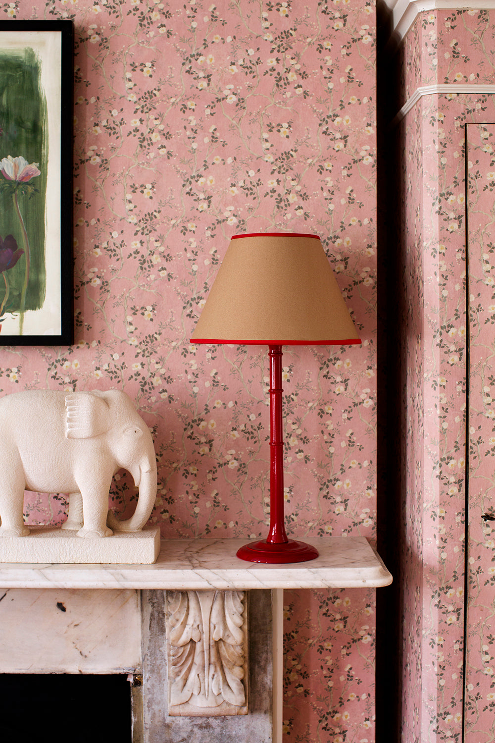A Rosi de Ruig brown paper lampshade with red cotton trim styled with a red glossy lamp on a mantlepiece